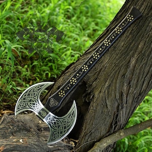 May include: A double-bladed axe with a black handle adorned with gold-colored star and vine designs. The silver-colored blades feature intricate Celtic knot patterns. The axe rests against a tree trunk, with a green leafy background.