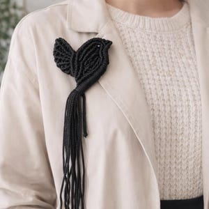 May include: A black woven bird brooch, featuring detailed wings and a long, trailing tail. The brooch is attached to a light-coloured jacket. The design creates a striking contrast.