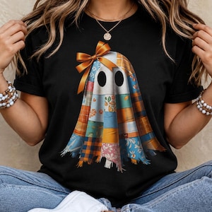 May include: A black t-shirt featuring a graphic of a ghost made of various colourful fabric patches, including plaid, floral, and autumn leaf patterns. The ghost has two black oval eyes and is adorned with a large, shiny orange ribbon bow on top.