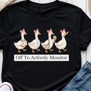 May include: Black t-shirt featuring a whimsical illustration of four geese, each with a pink bow and carrying various items. The text "Off To Actively Monitor" is displayed in a white rectangular box below the geese.