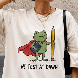 Puede incluir: Una camiseta de manga corta color hueso con un gráfico de una rana verde con capa roja, sosteniendo un libro de ciencias azul y un lápiz amarillo grande. Debajo de la rana, el texto dice WE TEST AT DAWN en una fuente negra sencilla.