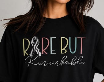 May include: A black short-sleeved t-shirt featuring the text "RARE BUT Remarkable" in pastel colors. The letter "A" in "RARE" is replaced by a black and white zebra-striped awareness ribbon. The word "Remarkable" is written in a white script font.