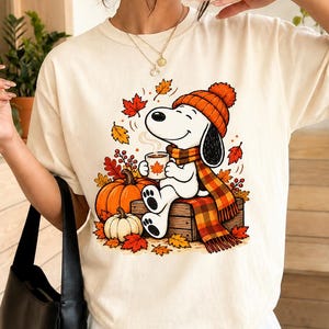 Peut inclure: Un t-shirt couleur crème arborant un motif de chien de dessin animé portant un bonnet en tricot orange et une écharpe à carreaux, assis sur une caisse en bois avec une tasse. Le design inclut des citrouilles orange et des feuilles d'automne.
