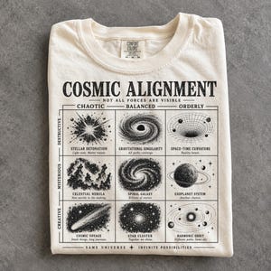 Könnte beinhalten: Ein cremefarbenes T-Shirt mit einem schwarzen Grafikdruck mit dem Titel COSMIC ALIGNMENT. Das Design zeigt ein Raster aus neun Illustrationen zum Thema Weltraum, unterteilt in chaotische, ausgeglichene und geordnete Kräfte.