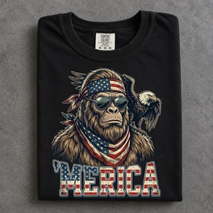 May include: A black crew neck t-shirt featuring a graphic of a brown sasquatch wearing a stars and stripes bandana and sunglasses, with a bald eagle perched on its shoulder. The text 'MERICA is printed below in a red, white, and blue flag pattern.