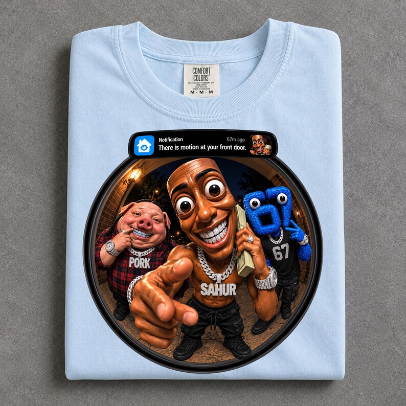 May include: A light blue t-shirt featuring a graphic print of a doorbell camera notification that reads "Notification 67m ago There is motion at your front door." The graphic shows a pig, a person, and a blue character with the number 67 on their shirt.