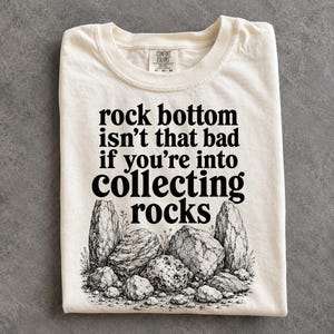 Pode incluir: Uma t-shirt de manga curta na cor creme com texto preto que diz: rock bottom isn't that bad if you're into collecting rocks. Abaixo do texto, há uma ilustração detalhada em preto e branco de várias rochas e pequenas plantas.
