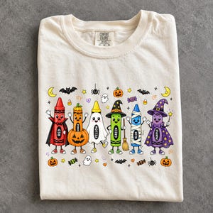 May include: A cream-coloured t-shirt featuring a graphic of six anthropomorphic crayons dressed in Halloween costumes. The crayons are labelled red, orange, yellow, green, blue, and purple, surrounded by small illustrations of bats, ghosts, pumpkins, and sweets.