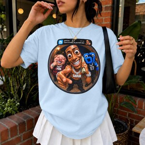 May include: A light blue t-shirt featuring a circular graphic print of a cartoon character with a large head, a pig in a red plaid shirt, and a blue number 67 character. The graphic includes a notification bar with the text "There is motion at your front door."