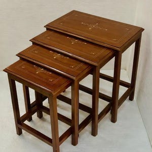 May include: A set of four wooden nesting tables in a rich brown hue. Each table has a rectangular top with inlaid decorative detailing. The tables are supported by square legs and are designed to be stored together.
