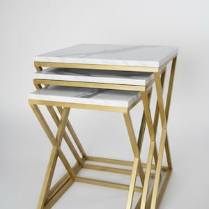 May include: A set of three nesting tables. Each table has a square top with a white marble effect and a gold metal frame. The tables are arranged in a nested configuration. The design is modern and stylish.
