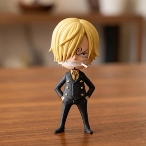 Puede incluir: Una figura de un personaje con cabello rubio y un traje negro. La figura tiene un cigarrillo en la boca y una sonrisa. El traje tiene botones dorados y una camisa dorada debajo. La figura está de pie con las manos en los bolsillos.