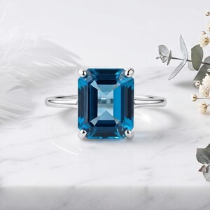 Pode incluir: Um anel de prata com uma grande pedra preciosa retangular azul-escura, de corte esmeralda, presa por quatro garras. A banda é fina e polida, apresentando um acabamento metálico limpo que complementa a cor azul profunda da pedra.