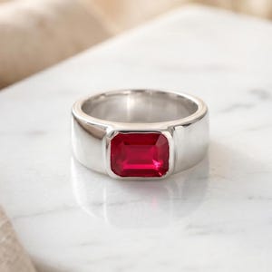 May include: A silver-toned metal ring featuring a single, rectangular, emerald-cut vibrant red gemstone set securely in the center. The band is smooth, polished, and has a wide, solid construction that tapers slightly toward the back of the ring.