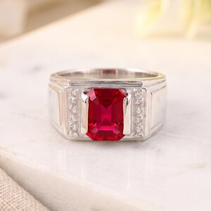 May include: A silver-toned ring featuring a large, rectangular, faceted red gemstone set in the center. The band is flanked by two vertical rows of small, clear, round stones on either side of the central red stone, creating a structured design.