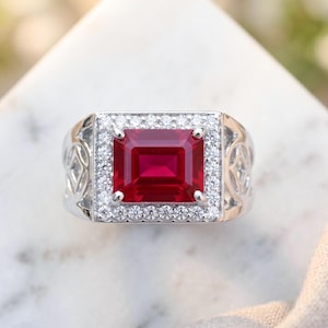 May include: A silver-toned ring featuring a large, rectangular, deep red gemstone at the center. The stone is surrounded by a halo of small, clear, round-cut stones. The band has an intricate, open-work metal design on the sides of the setting.
