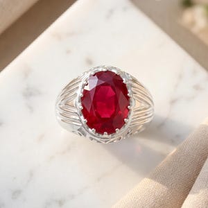 Peut inclure: Une bague en argent ornée d'une grande pierre précieuse rouge foncé de taille ovale, fixée par des griffes. L'anneau de la bague présente un motif nervuré et texturé qui s'étend du sertissage central le long des côtés de la bande métallique.
