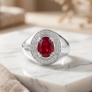 May include: A silver-toned ring featuring a central oval-cut deep red ruby gemstone. The ruby is surrounded by a halo of small, clear, round-cut white stones set into the metal band, creating a bright, reflective, and polished appearance.