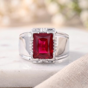 May include: A silver-toned ring featuring a large, rectangular, faceted red gemstone set in the center. The metal band has a polished finish with vertical grooved detailing flanking the stone on both the top and bottom edges of the setting.
