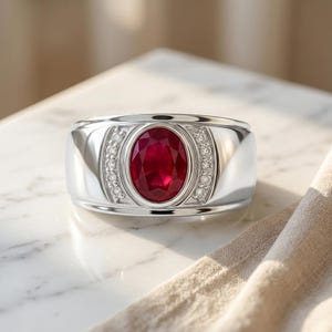 May include: A polished silver-tone ring featuring a large, oval-cut deep red ruby at the centre. The ruby is framed by two vertical rows of small, clear round-cut stones set into the band on either side of the central gemstone setting.