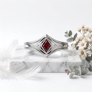 Puede incluir: Un brazalete de metal plateado con una piedra preciosa de color rojo intenso tallada en forma de marquesa en el centro. La banda presenta un diseño geométrico en capas con líneas grabadas y detalles puntiagudos alrededor de la piedra.