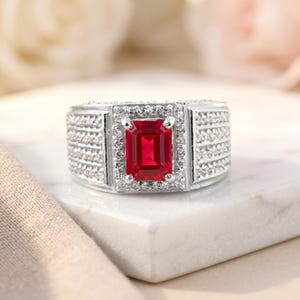 May include: A silver-toned ring featuring a central emerald-cut red gemstone surrounded by a halo of small white stones. The wide band is decorated with three rows of tiny white stones on each side, creating a textured and sparkling appearance.