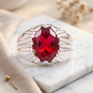 May include: A silver ring featuring a large, oval-cut, deep red gemstone set in the center. The metal band has a polished finish with decorative, radiating grooves extending from the central stone setting toward the sides of the ring.