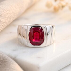 May include: A silver-toned ring featuring a large, cushion-cut, vibrant red gemstone set in the center. The band is wide and polished, characterized by vertical grooved lines flanking the central stone setting, creating a structured appearance.