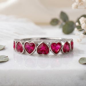 May include: A silver ring with a row of heart-shaped, bright red gemstones. The gemstones are set closely together, forming a continuous pattern around the band. The ring is presented on a white surface, with a soft, blurred background.
