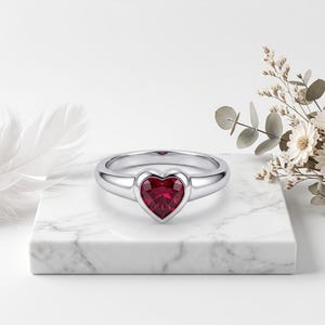 Pode incluir: Um anel de tom prateado com uma pedra preciosa vermelha central em forma de coração, engastada num bisel liso. A banda é polida e arredondada, criando uma superfície limpa e refletora. O design destaca o coração vermelho vibrante.