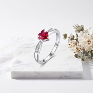 May include: A silver-toned ring featuring a central heart-shaped red gemstone held in a prong setting. The band is accented with small, clear stones embedded along the shoulders, which are framed by a delicate milgrain edge detail.