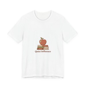 May include: White t-shirt featuring a graphic of a red apple atop a stack of books, with the text "Quiet Influence" below. The design is in shades of red, brown, and green. The shirt is a casual wear item.