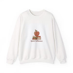 May include: White sweatshirt with a graphic of a red apple atop a stack of books. The text "Quiet Influence" is printed below the books in brown. The sweatshirt has a crew neck and long sleeves.