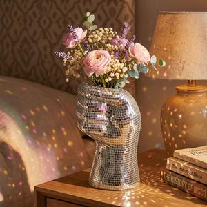 May include: A silver mosaic vase in the shape of a clenched fist, holding pink roses, lavender, and foliage. The vase is on a wooden bedside table, next to a lamp and books. The vase is approximately 20 cm tall.