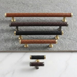 Puede incluir: Tiradores de armario con asas trenzadas de imitación cuero en varios colores, incluyendo marrón, negro y beige. Los tiradores tienen detalles en latón o metal oscuro. El herraje está diseñado para muebles y armarios.