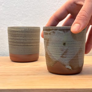 Op de afbeelding: Twee handgemaakte keramische bekers. Eén is scherp, vastgehouden door een hand, en de andere is enigszins onscherp. Beide hebben een bruine basis en een getextureerd, lichtgroen en bruin bovenstuk. De bekers staan op een houten oppervlak.