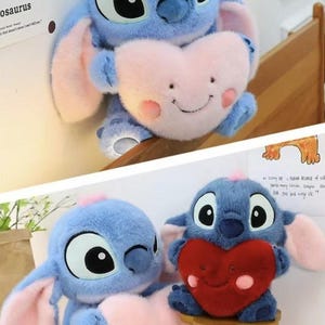 May include: Blue plush toy resembling a cartoon character, holding a pink heart-shaped pillow with a smiling face. The toy has large black eyes, pink ears, and a small pink tuft of hair. The heart pillow has rosy cheeks.