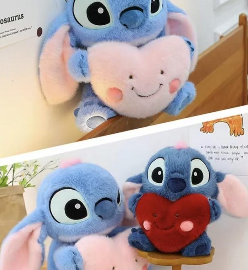 May include: Blue plush toy resembling a cartoon character, holding a pink heart-shaped pillow with a smiling face. The toy has large black eyes, pink ears, and a small pink tuft of hair. The heart pillow has rosy cheeks.