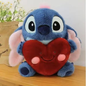 May include: A plush blue character with large black eyes, pink ears, and a pink tuft on its head, holding a red heart-shaped pillow with a smiling face. The plush toy is sitting on a wooden surface.