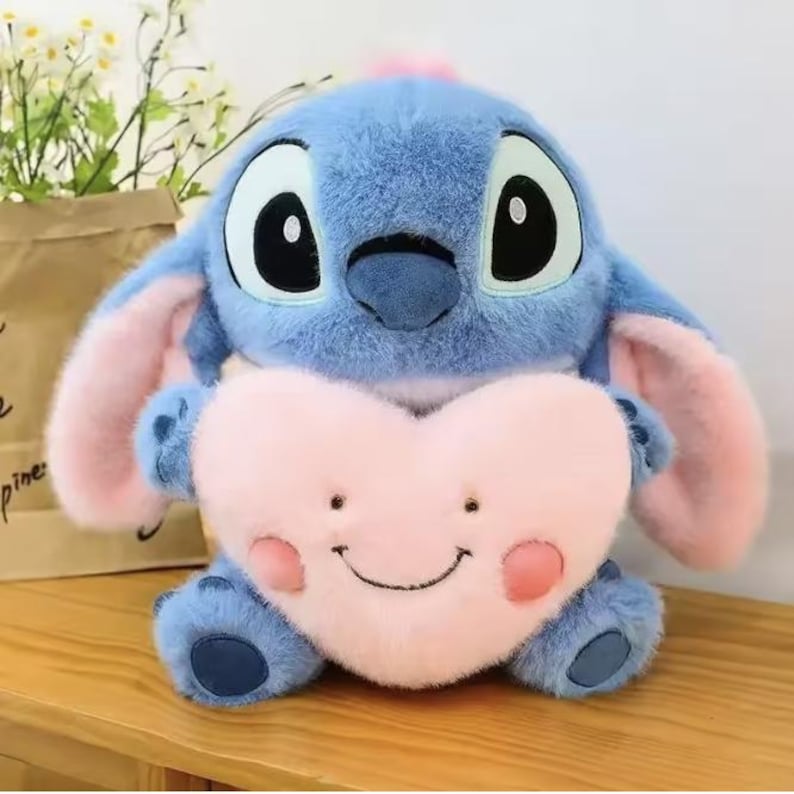 May include: A blue plush toy with large black eyes, pink ears, and a pink heart-shaped pillow. The plush toy is holding the pillow, which has a smiling face and pink cheeks.