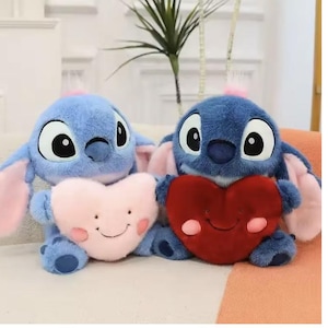 May include: Two blue plush toys, each resembling a cartoon character, holding heart-shaped pillows. One pillow is pink with a smiling face, the other is red. The toys have large eyes and pink accents.
