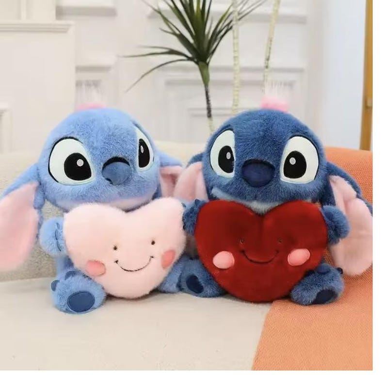 May include: Two blue plush toys, each resembling a cartoon character, holding heart-shaped pillows. One pillow is pink with a smiling face, the other is red. The toys have large eyes and pink accents.