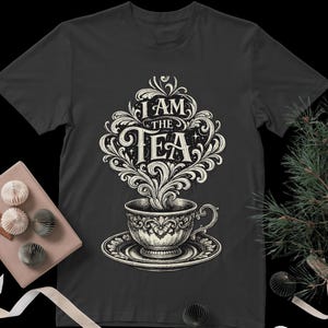 Può includere: T-shirt grigio scuro con un'illustrazione dettagliata di una tazza da tè e un piattino. Il vapore sale a formare le parole "I AM THE TEA" in un design ornato. Il design è nei colori bianco e crema.
