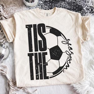 Peut inclure: T-shirt cr&egrave;me avec l'inscription noire "TIS THE SEASON", accompagn&eacute;e d'un graphisme de ballon de football noir et blanc. Le t-shirt a des manches courtes et un col rond.