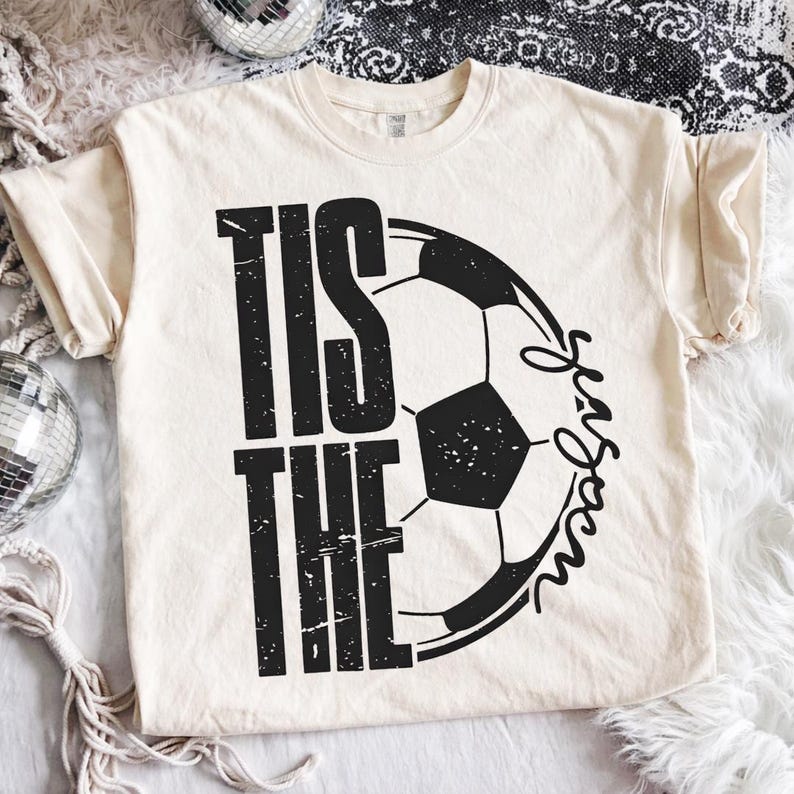 Peut inclure: T-shirt cr&egrave;me avec l'inscription noire "TIS THE SEASON", accompagn&eacute;e d'un graphisme de ballon de football noir et blanc. Le t-shirt a des manches courtes et un col rond.