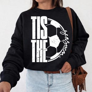 Peut inclure: Sweat-shirt noir avec l'inscription blanche "TIS THE SEASON" et un motif de ballon de football. La personne porte un jean bleu clair et un sac marron.