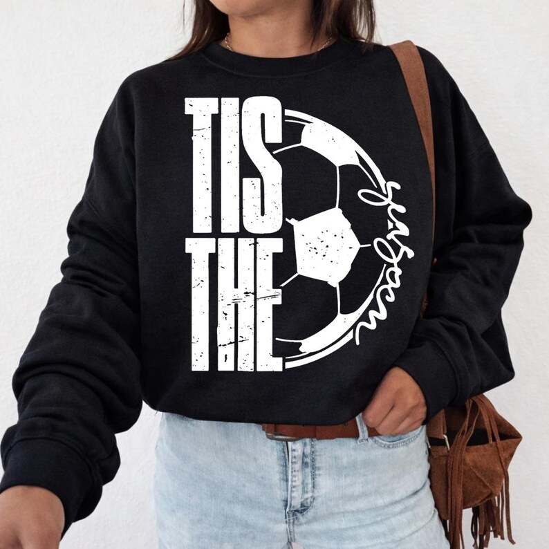 Peut inclure: Sweat-shirt noir avec l'inscription blanche "TIS THE SEASON" et un motif de ballon de football. La personne porte un jean bleu clair et un sac marron.