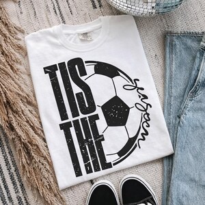 Peut inclure: T-shirt blanc avec un motif de ballon de football noir et le texte "Tis the Season". Le t-shirt est associ&eacute; &agrave; un jean bleu et des baskets noires et blanches. Le motif est centr&eacute; sur le devant.