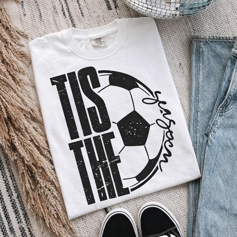 Peut inclure: T-shirt blanc avec un motif de ballon de football noir et le texte "Tis the Season". Le t-shirt est associ&eacute; &agrave; un jean bleu et des baskets noires et blanches. Le motif est centr&eacute; sur le devant.