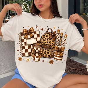 Pode incluir: Uma t-shirt branca com uma estampa gráfica de um caderno xadrez castanho e branco, uma mochila com padrão de leopardo, um copo de café e lápis num copo. Cada item é decorado com laços castanhos e o design inclui pequenos detalhes de estrelas e flores.